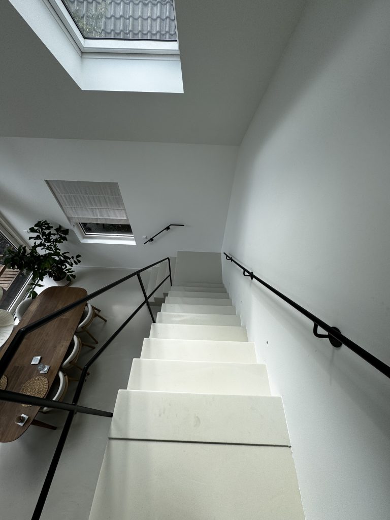 Moderne witte trap met zwarte leuning, veel natuurlijk licht en elegante, minimalistische uitstraling.